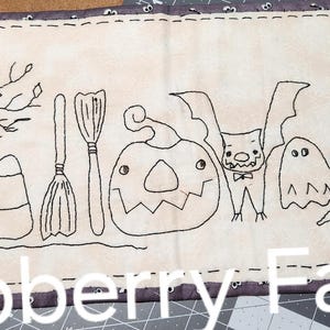 May include: A rectangular, beige Halloween-themed fabric banner with black embroidered designs. The banner features a tree with an owl, candy corn, a broom, a jack-o'-lantern, a bat, ghosts, and a witch's hat. The words "Wildoberry Farms" are at the bottom.