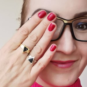 Pode incluir: Uma mulher vestindo um suéter de gola alta rosa e óculos com armação dourada. Ela está usando dois anéis em seu dedo, ambos com pedras pretas em forma de triângulo.