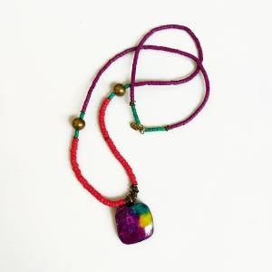 Peut inclure: Un collier multicolore avec un pendentif carré. Le collier est composé de perles rouges, violettes et vertes. Le pendentif est un carré de couleur violette, verte et jaune.