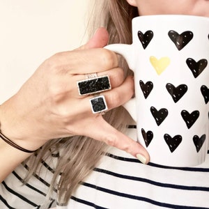 Könnte beinhalten: Zwei schwarze rechteckige Ringe mit einer strukturierten Oberfläche. Die Ringe werden am Zeige- und Mittelfinger einer Hand getragen, die eine weiße Tasse mit schwarzen Herzen und einem einzelnen gelben Herzen hält.