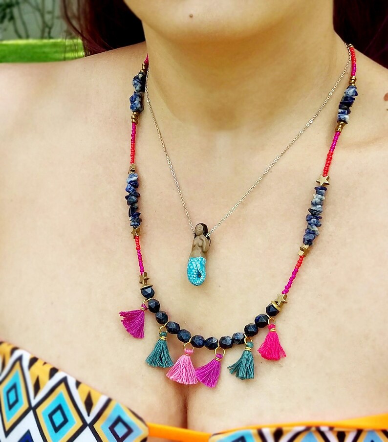 May include: A layered necklace with a mermaid charm pendant. The necklace features blue and black beads, pink and green tassels, and gold star accents.