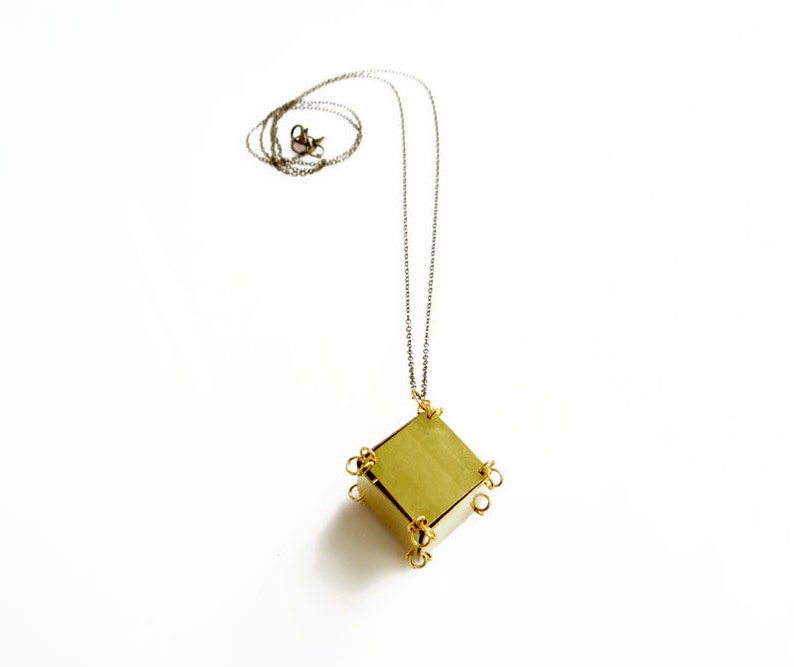 May include: A gold necklace with a geometric pendant. The pendant is a cube with a square face and is made of brass. The necklace is on a white background.