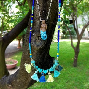 May include: A turquoise and blue beaded necklace with a mermaid pendant and turquoise stone accents. The necklace has yellow and blue tassels.
