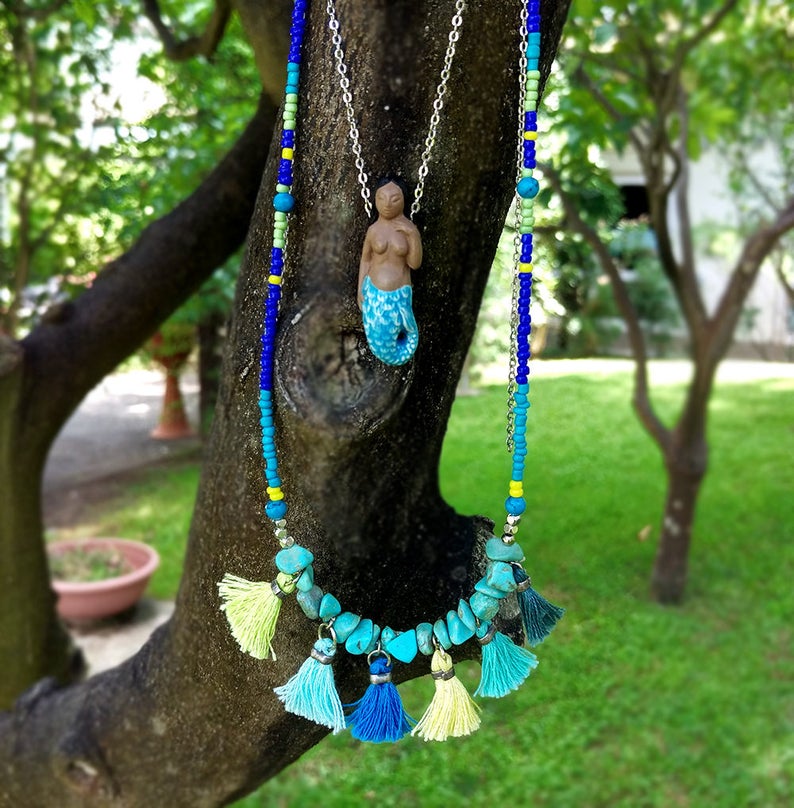 May include: A turquoise and blue beaded necklace with a mermaid pendant and turquoise stone accents. The necklace has yellow and blue tassels.