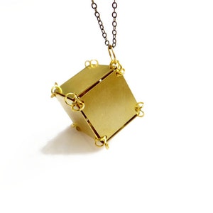 May include: A gold-toned pendant necklace with a geometric cube-shaped charm. The cube is made of multiple pieces of metal that are connected by small rings. The necklace is hanging from a simple chain.