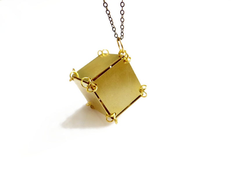 May include: A gold-toned pendant necklace with a geometric cube-shaped charm. The cube is made of multiple pieces of metal that are connected by small rings. The necklace is hanging from a simple chain.