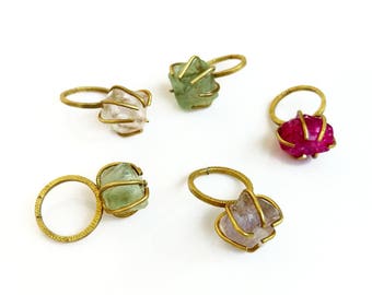 May include: Five brass rings with different colored gemstones. Each ring features a unique, geometric design.