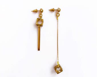 May include: A pair of gold earrings with a geometric design. One earring features a square with a circle cutout and a long, thin bar hanging from it. The other earring has a square with a circle cutout hanging from a chain.