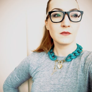 May include: A woman wearing black framed glasses and a teal blue chain necklace with a gold heart-shaped lock and key pendant.