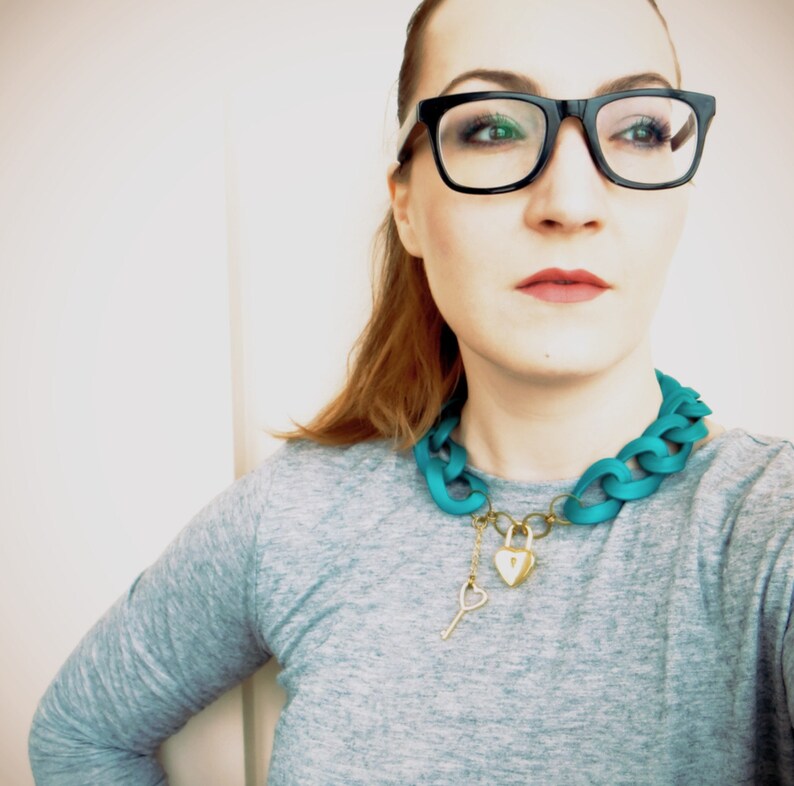 May include: A woman wearing black framed glasses and a teal blue chain necklace with a gold heart-shaped lock and key pendant.