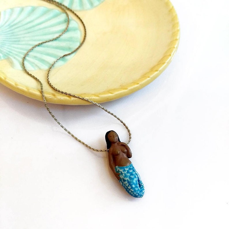 May include: A silver chain necklace with a small, hand-crafted mermaid pendant. The mermaid is brown-skinned with blue and white scales and is posed sitting with her hand on her chin.