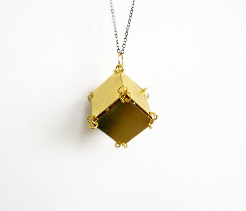May include: A gold-toned necklace with a geometric pendant made of multiple brass squares. The pendant is a cube shape with a chain attached to the top.