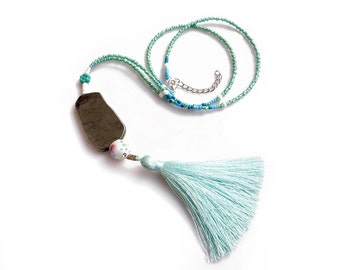 Puede incluir: Un collar hecho a mano con un colgante rectangular gris oscuro. El collar tiene una cadena de cuentas en tonos verdes y azules, con una borla azul claro. Una cuenta blanca con un patrón floral añade detalles.