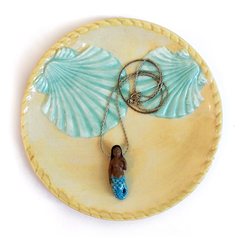 May include: A ceramic dish featuring two blue seashells and a gold chain necklace with a mermaid pendant. The mermaid is brown with a blue tail.