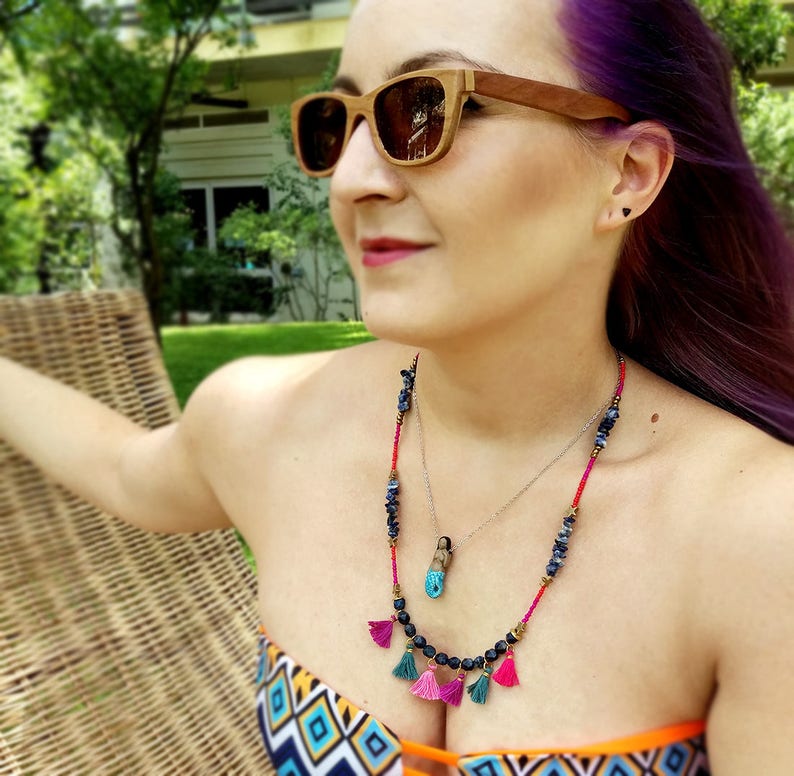 May include: A woman wearing a colourful bikini top with a layered necklace featuring blue and black beads, a small pendant, and pink and turquoise tassels.
