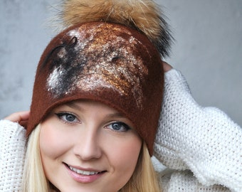 May include: A woman wearing a brown felt hat with a gold and black abstract design and a fluffy pom pom. The hat is worn over a white cable knit sweater.