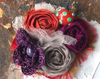 May include: Close-up of a mixed media artwork featuring fabric flowers and a glass bead. The flowers are in shades of red, purple, and gray, with a white lace accent. A round, orange glass bead with green dots sits atop the arrangement.