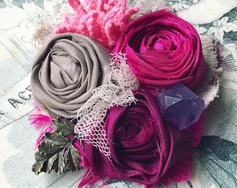 May include: A handmade fabric flower brooch with three pink and gray fabric roses, a purple crystal, and a bronze leaf accent.