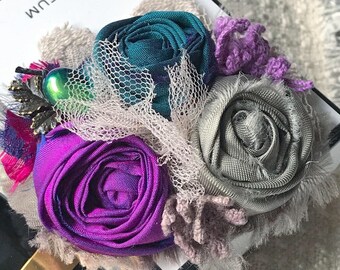 May include: A handmade fabric flower brooch with teal, purple, and gray fabric flowers. The brooch is decorated with lace and a small metal charm. The brooch is sitting on a white box with the word "PARFUM" printed on it.