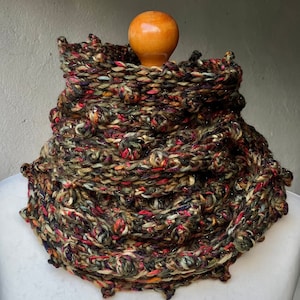 May include: A multi-colored crocheted cowl with a textured, bumpy surface. The cowl is made with a variety of yarns in shades of green, brown, red, and orange. The cowl is displayed on a white mannequin.