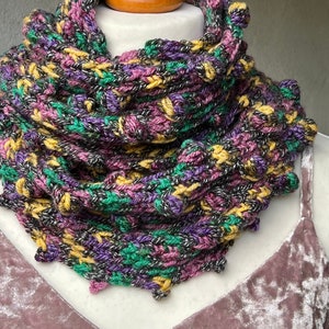 May include: A colorful crocheted cowl scarf in shades of purple, green, yellow, and black. The scarf is made with a textured stitch and has a slightly ruffled edge.