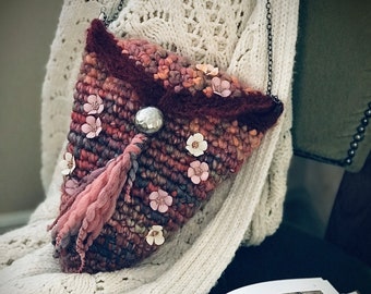 May include: A small, triangular, woven bag with a pink tassel and floral embellishments. The bag is made of a variety of colors, including brown, pink, and red. The bag is hanging from a silver chain.