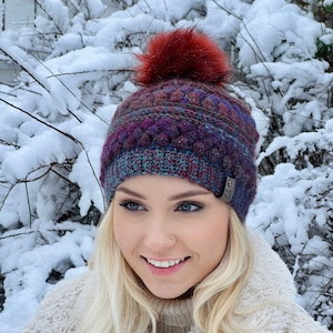 Puede incluir: Una mujer lleva un gorro de punto morado, azul y rojo con un pompón burdeos. El gorro tiene un diseño trenzado y una pequeña etiqueta plateada con la palabra "No" en ella.