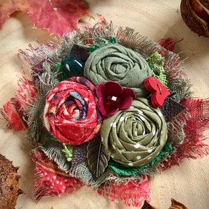 May include: A handmade fabric flower brooch with red, green, and brown fabric flowers. The brooch is decorated with a green gem, a red gem, and a brown leaf.