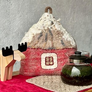 以下が含まれることがあります： 雪に覆われた屋根の家型をした赤いフェルトティーコージー。黒い蓋のガラスティーポットと木製のヘラジカの置物も写っています。コージーには小さな窓とループハンドルが付いています。