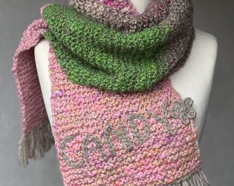 Può includere: Una sciarpa di lana rosa, verde e marrone con frange. La sciarpa ha la parola "Candy" ricamata in filo grigio.