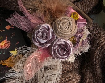 May include: A delicate floral brooch with three fabric roses in shades of purple and beige, accented with a pink tulle bow and a feather. The brooch is set against a brown knitted fabric background.