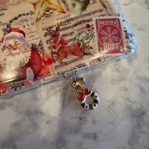 May include: A clear phone case with a Christmas theme. The case features a collage of vintage images, including Santa Claus, a reindeer, and a star. A gold charm with a wreath and a red bow hangs from the case.