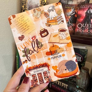 May include: A rectangular sticker with an orange and white plaid pattern, featuring fall-themed illustrations and text. Designs include a ghost, pumpkins, a bookstore, and the words "I love fall" and "hello pumpkin". The sticker is held in a hand.