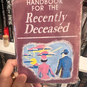 Puede incluir: Un libro de color burdeos titulado "Handbook for the Recently Deceased". La portada presenta una ilustraci&oacute;n de una pareja con atuendo rosa y azul, sobre un cielo con nubes y un sol amarillo. El libro est&aacute; en una funda protectora transparente.