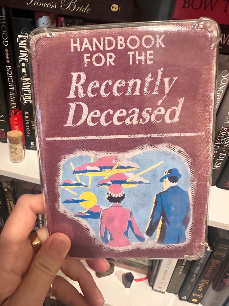 Puede incluir: Un libro de color burdeos titulado "Handbook for the Recently Deceased". La portada presenta una ilustraci&oacute;n de una pareja con atuendo rosa y azul, sobre un cielo con nubes y un sol amarillo. El libro est&aacute; en una funda protectora transparente.