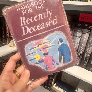 Puede incluir: Un libro titulado "Handbook for the Recently Deceased" con una cubierta burdeos y una funda protectora transparente. La portada presenta una ilustraci&oacute;n retro de dos figuras bajo un cielo estilizado. El libro se sostiene frente a una estanter&iacute;a.