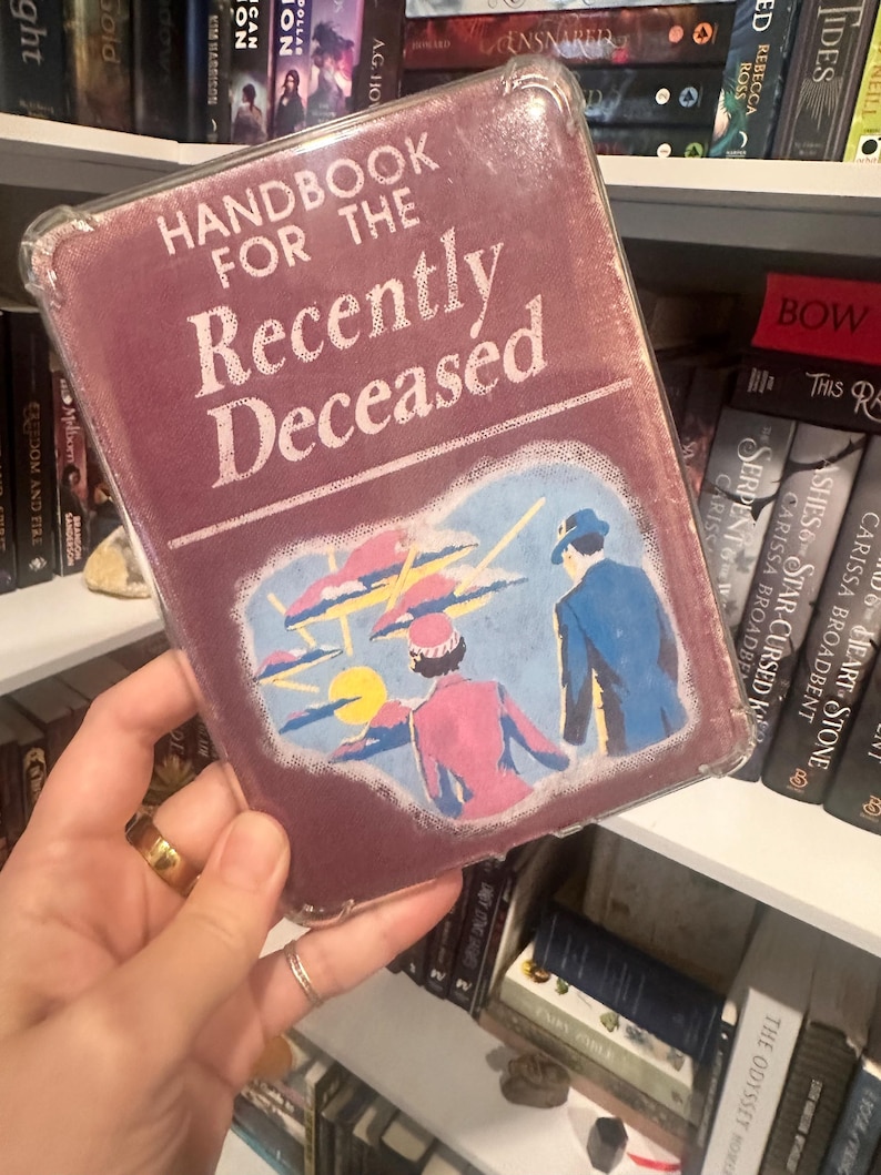 Puede incluir: Un libro titulado "Handbook for the Recently Deceased" con una cubierta burdeos y una funda protectora transparente. La portada presenta una ilustraci&oacute;n retro de dos figuras bajo un cielo estilizado. El libro se sostiene frente a una estanter&iacute;a.