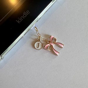 Op de afbeelding: Een goudkleurige bedel met een roze strik en een witte ronde hanger met de letter "J", bevestigd aan een Kindle. De strik heeft roze emaille en gouden randen. De Kindle zit in een doorzichtige hoes.