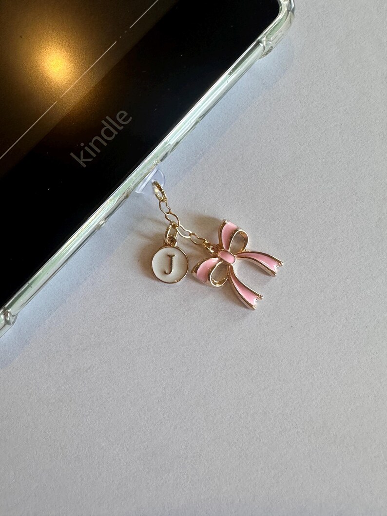Op de afbeelding: Een goudkleurige bedel met een roze strik en een witte ronde hanger met de letter "J", bevestigd aan een Kindle. De strik heeft roze emaille en gouden randen. De Kindle zit in een doorzichtige hoes.