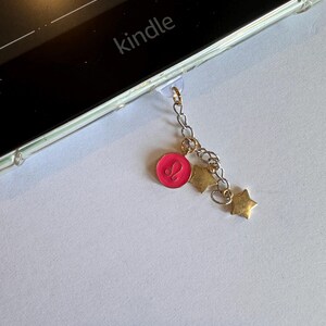 Op de afbeelding: Een bedel voor een Kindle e-reader met een roze dierenriemteken hanger en gouden ster bedels. De bedel is bevestigd aan de Kindle met een doorzichtige plastic lus. Het woord "kindle" is zichtbaar.