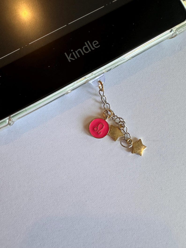 Op de afbeelding: Een bedel voor een Kindle e-reader met een roze dierenriemteken hanger en gouden ster bedels. De bedel is bevestigd aan de Kindle met een doorzichtige plastic lus. Het woord "kindle" is zichtbaar.