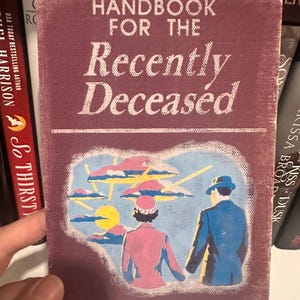 Puede incluir: Un libro titulado "Handbook for the Recently Deceased" con una portada burdeos. La portada presenta una ilustraci&oacute;n de dos personas en un paisaje con un sol amarillo y cielo azul. Se ven otros libros.