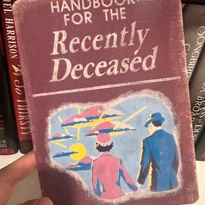 Puede incluir: Una portada de libro de estilo vintage con el t&iacute;tulo "Handbook for the Recently Deceased" en letras blancas sobre un fondo granate. La portada presenta una ilustraci&oacute;n de dos figuras en un paisaje surrealista.