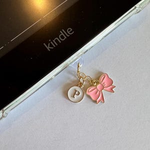 Op de afbeelding: Een goudkleurige bedel met een roze strik en een witte cirkel met de letter "P", bevestigd aan een doorzichtige Kindle-hoes. Het woord "kindle" is zichtbaar op het apparaat.