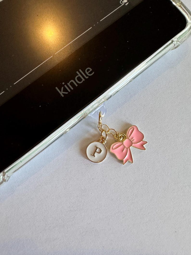 Op de afbeelding: Een goudkleurige bedel met een roze strik en een witte cirkel met de letter "P", bevestigd aan een doorzichtige Kindle-hoes. Het woord "kindle" is zichtbaar op het apparaat.