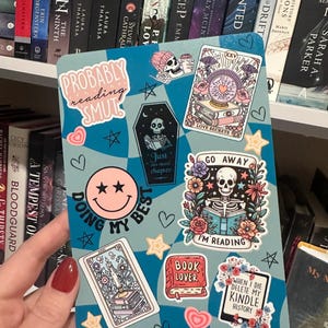 May include: A blue and white sticker with a checkered pattern and various stickers on top. The stickers feature illustrations of skeletons, flowers, hearts, stars, and text that reads "Probably reading smut", "Just one chapter", "Go away", "Doing my best", "I'm reading", "Book lover", and "When I die delete my kindle history".
