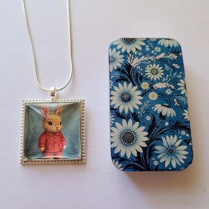 May include: A silver necklace with a square pendant featuring a cartoon bunny wearing a red sweater. The pendant is set against a blue background. The necklace comes in a small blue tin with a white floral pattern.