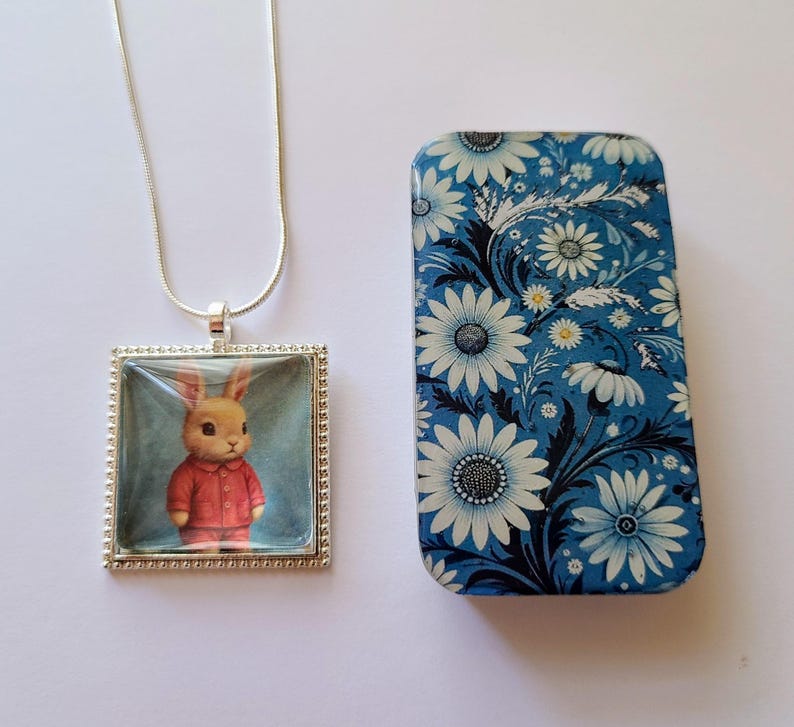 May include: A silver necklace with a square pendant featuring a cartoon bunny wearing a red sweater. The pendant is set against a blue background. The necklace comes in a small blue tin with a white floral pattern.