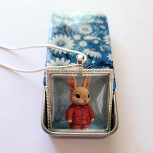 May include: A silver necklace with a square glass pendant featuring a cartoon bunny wearing a red jumpsuit. The pendant is surrounded by a silver frame with a beaded border.