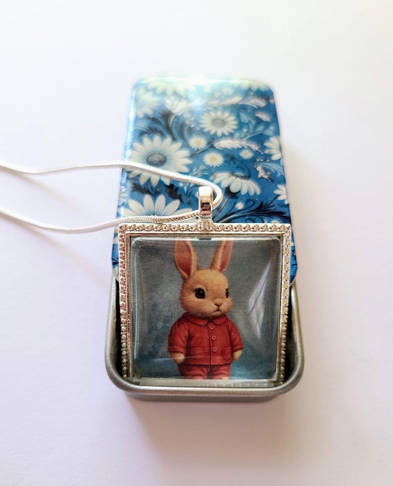 May include: A silver necklace with a square glass pendant featuring a cartoon bunny wearing a red jumpsuit. The pendant is surrounded by a silver frame with a beaded border.